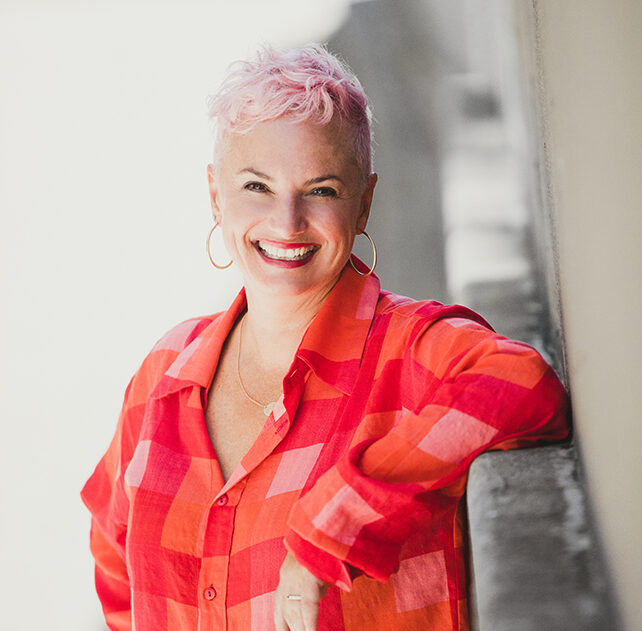Image of Lucy Suze with short pink hair leaning against a wall.